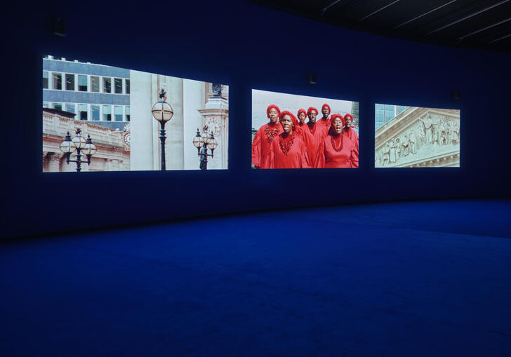 Still image of blue gallery floor and 3 screens on the wall depicting Julianknxx work