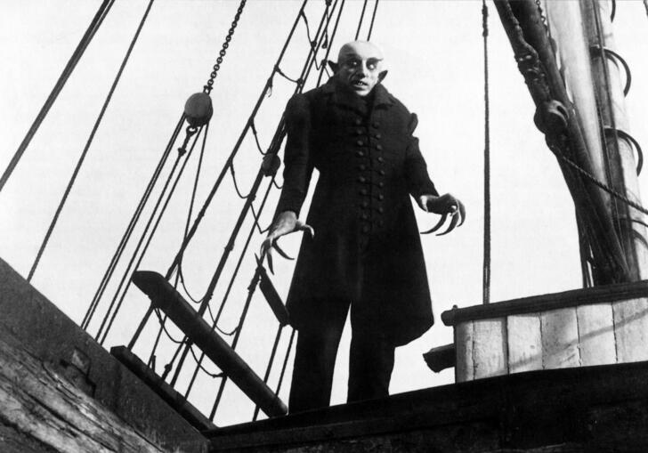 Still from 1922 silent film Nosferatu: A Symphony of Horror, showing Nosferatu with his long, claw-like fingers and pointed ears, standing on a ship wearing a long black coat