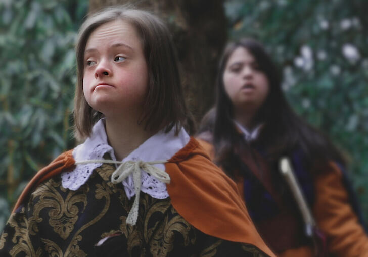 Two young girls in period dress stand stoically in a forest. 