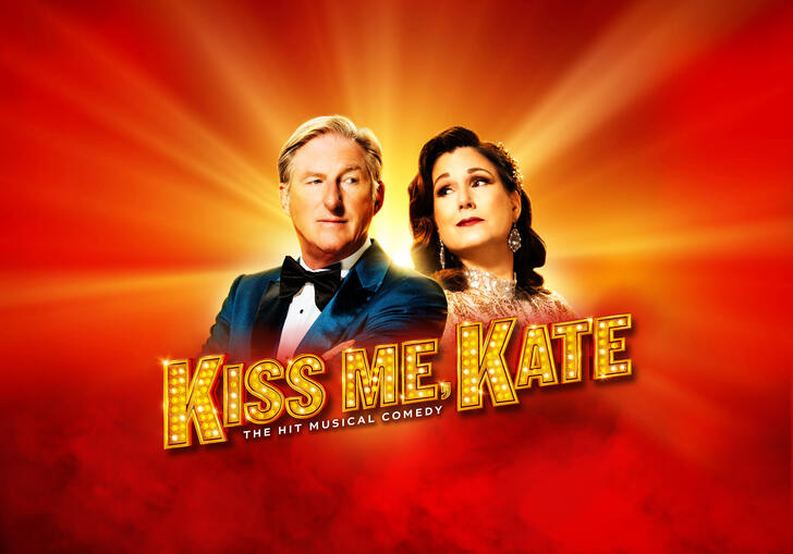 Adrian Dunbar and Stephanie J. Block stand side-by-side against a red background which has a yellow glow. The words 'Kiss Me, Kate, The Hit Musical Comedy' is written across it.