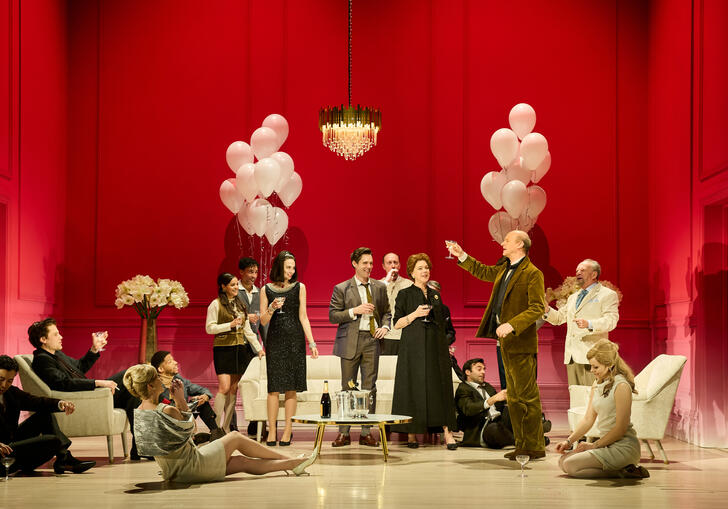 A group of people sit on stage at a party scene, around a large table. 