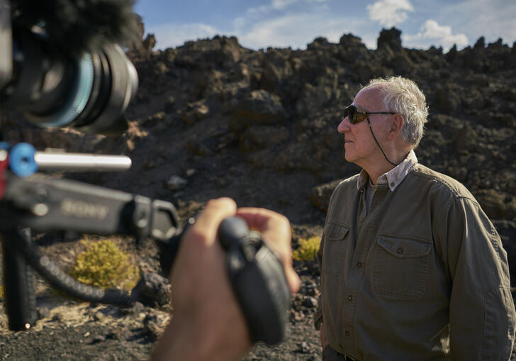Werner Herzog stands on a tree covered rural landscape, being filmed by a camera.