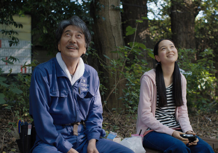 A man and a girl sit on a bench in a park, looking up to the sky and smiling. 