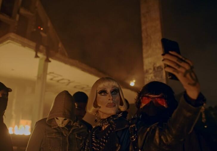 A woman with heavy makeup takes a selfie in front of a burning building, surrounded by masked people.