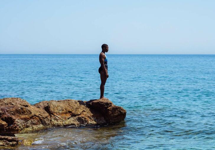 A woman in a bathing suit stands on a rock, looking out across the blue ocean in front of her.