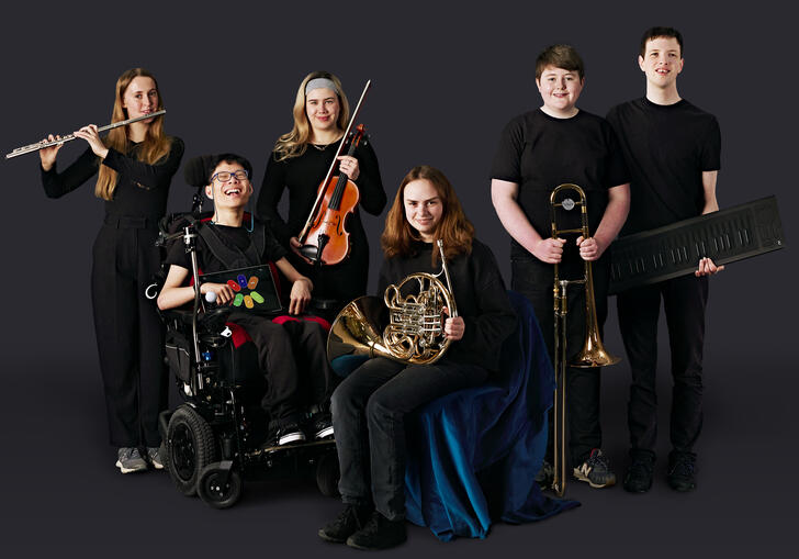 Musicians from the National Open Youth Orchestra with their instruments, in front of a dark grey background.