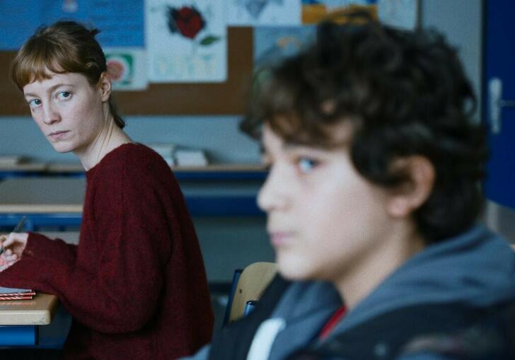 A teacher looks across the desks at a young boy student. 