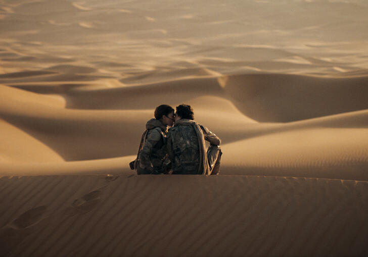 Two young people sit atop a sand dune in the vast expanse of the empty desert.