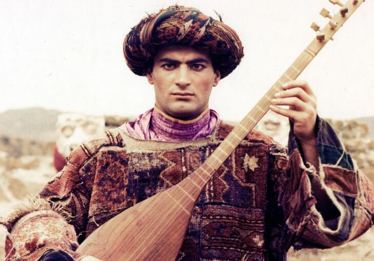 A young person playing a lute in traditional clothing, against a desert scape.