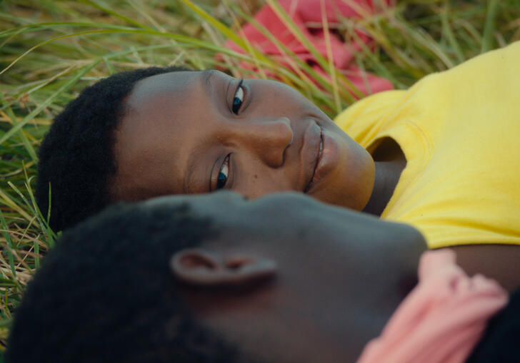 A young couple lie in tall grass, gazing into each other's eyes. 