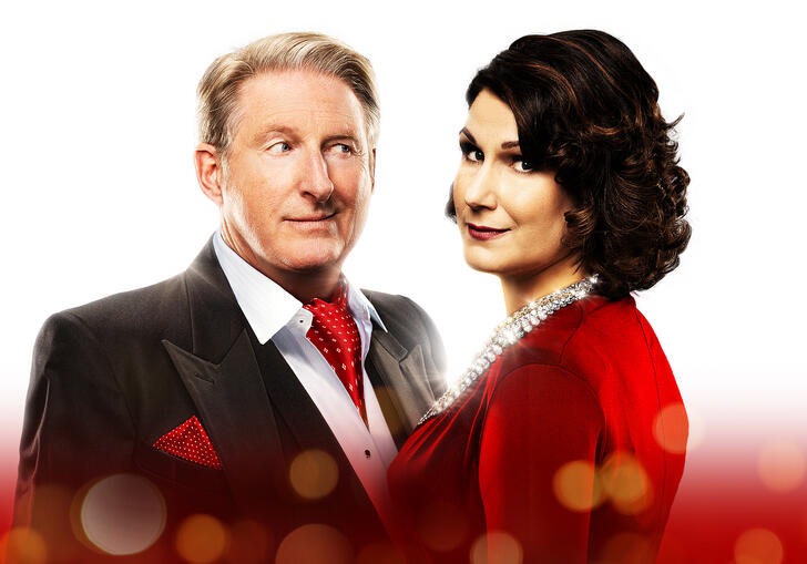 Adrian Dunbar and Stephanie J. Block pose alongside each other against a white and red background. Stephanie smiles down the camera, while Adrian looks at her cheekily. 