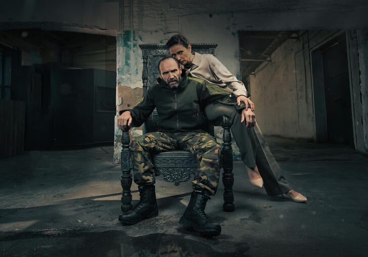 A man wearing army gear sits on a throne with a woman leaning over him. 