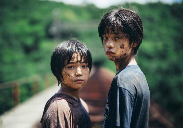 Two young boys covered in mud stand on train tracks in a rural setting, looking towards the camera.