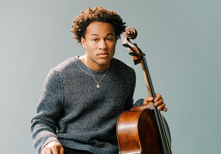 Sheku Kanneh-Mason sitting on a wooden stool with his cello, in front of a pale blueish gray background