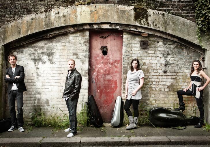 The Carducci Quartet standing in a line in front of an old, dilapidated building, their instruments in cases on the floor beside them.