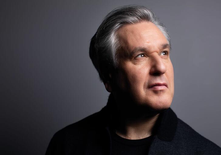 A portrait photo of Sir Antonio Pappano, against a grey background, looking away from the camera into the distance.