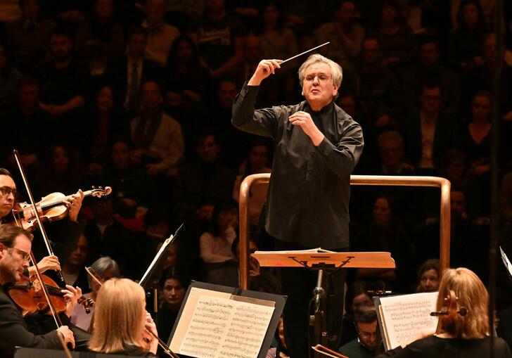 Sir Antonio Pappano conducting the LSO on the Barbican stage, holding a baton, with LSO string players in the foreground of the image and audience members in the background.