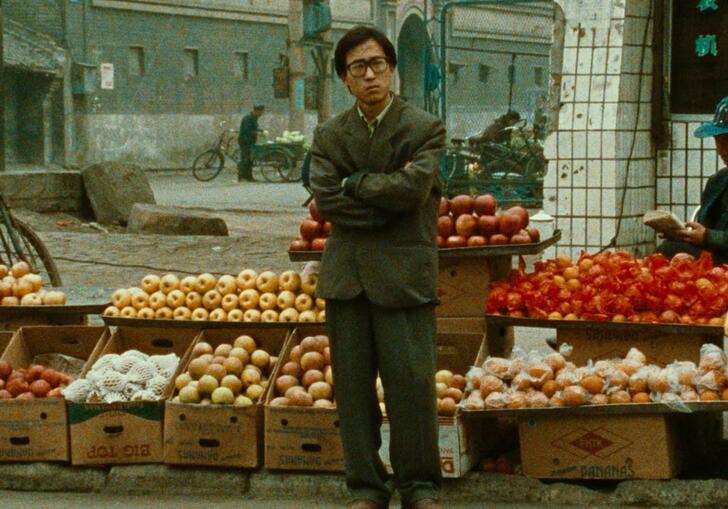 A man stands in front of a fruit stall with his arms folded across his chest.