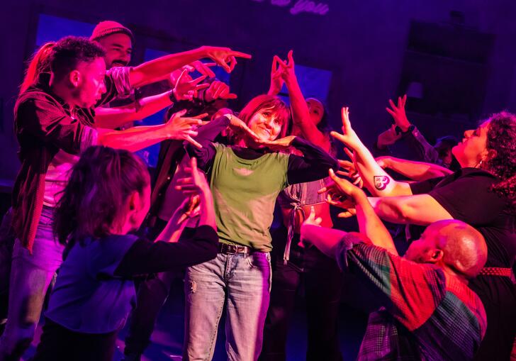 A group of performers gather around one performer and raise their hands towards her.
