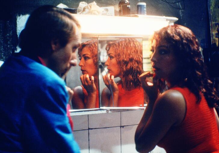 Two people stand in front of a mirror in a darkly lit bathroom. 