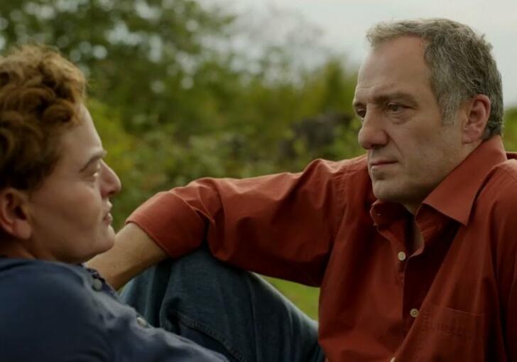 A middle-aged woman in a blue dress is sat on grass staring into the eyes of a man in a red shirt.