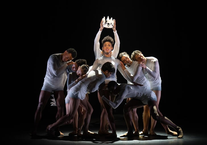 a group of ballerinas crouch a person in the centre holding a crown over their head
