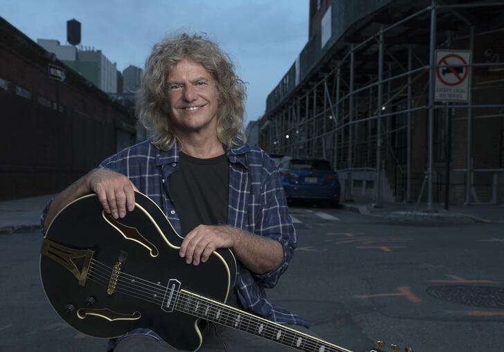 Pat Metheny sits, holding his guitar, on the street. 