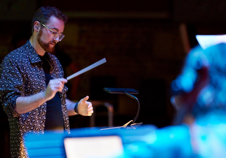 Composer Anselm McDonnell conducing with a baton. He has a colourful patterned shirt, glasses and red hair.