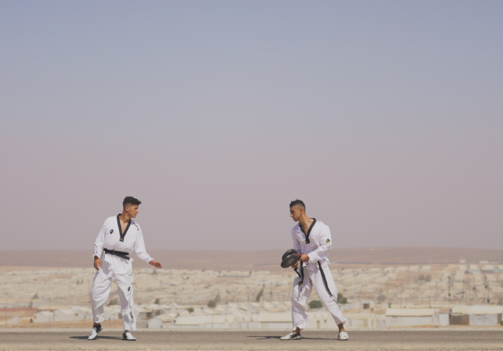 Two boys stand opposite each other wearing mixed martial arts uniforms, in front of a desert city. 