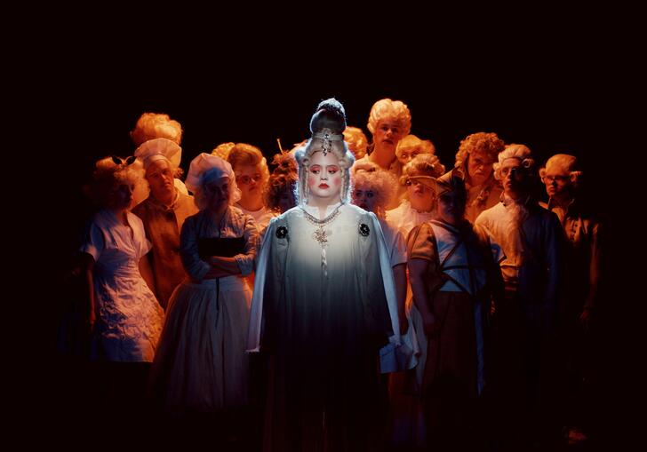 A group of people in theatrical period dress stand in a circle in a darkly lit space.