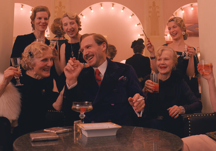 A hotelier sits smoking and laughing, surrounded by glamorous older women, in a nice looking hotel salon.