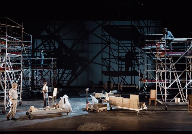 Two people stand on stage with a minimal set of furniture while tall scaffold towers line the edge of the stage.
