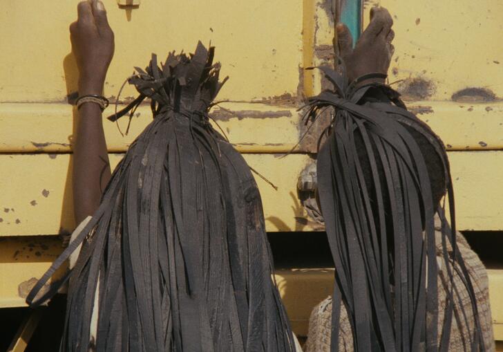 Two men with black rubber headdresses stand in front of a yellow truck. 