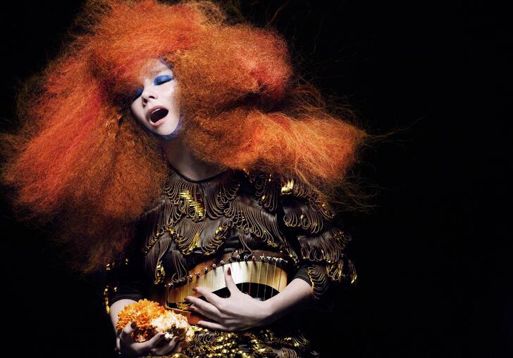 Bjork, large red hair in full costume bends over a dark background. 