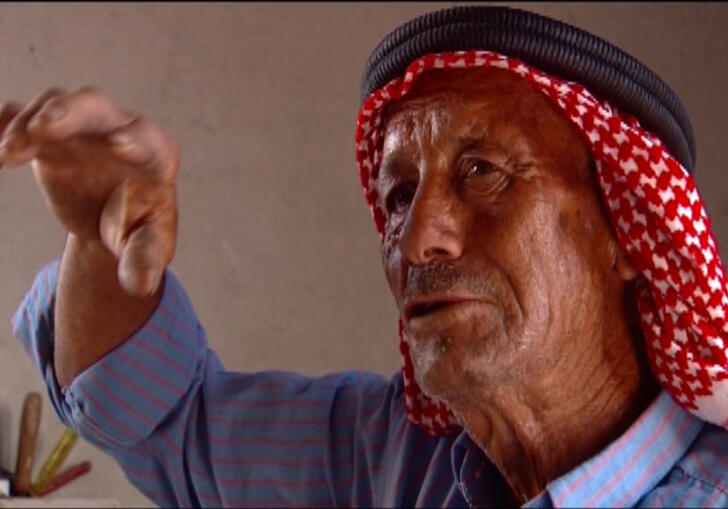 An older man in a blue shirt and head wrap talks whilst animating with his hand.