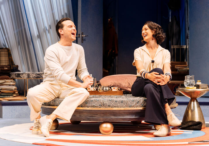 A man and a woman sit on a divan on stage laughing with each other. 