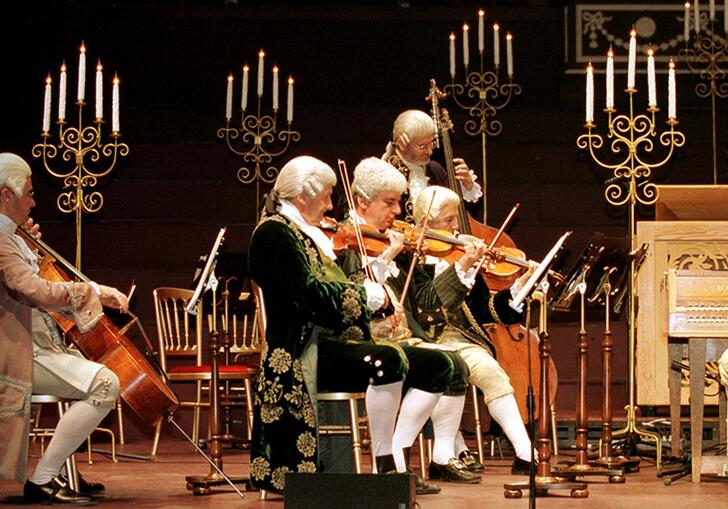 Musicians performing on stage in period costume, with five tall candelabras behind them