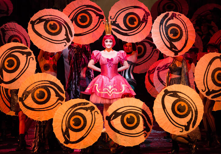 An actor stands in the middle of the stage in a pink dress, surrounded by large eyes. 