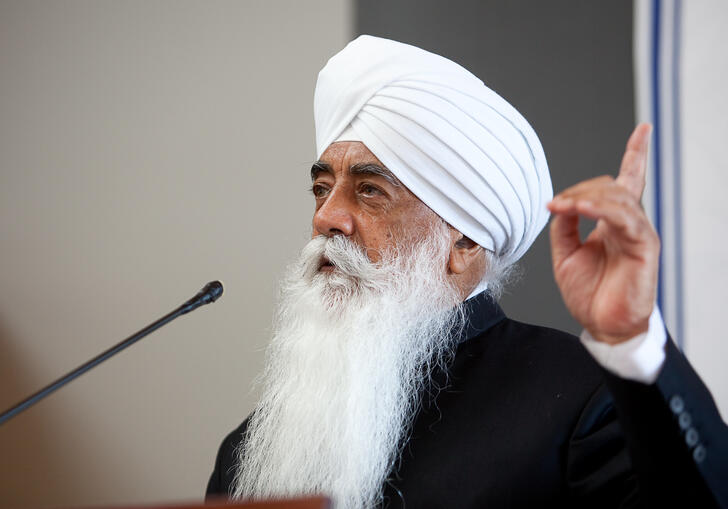 Bhai Sahib Mohinder Singh Ji speaking into a microphone, holding his finger up