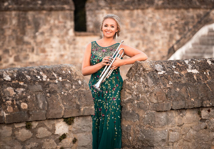 Matilda Lloyd in a green dress, holding her trumpet. She is stood within the walls of an old stone castle.