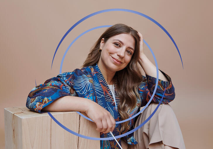Dalia Stasevska sitting down, resting her arm on a wooden block. There are blue circular swirls around her.