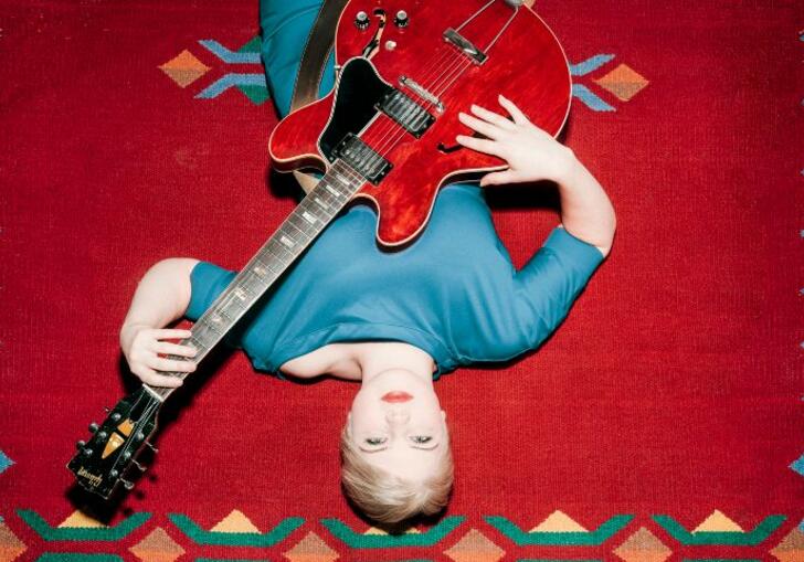 LILJA lays on a red and blue carpet, holding a red guitar