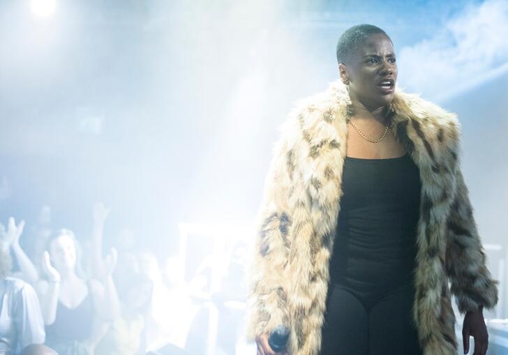 A person in a fur coat stands on stage as people dance in the background in a dark room lit by haze.