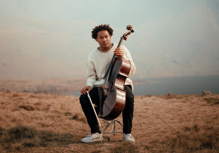 Sheku Kanneh-Mason sitting with his cello in a mountainous landscape, with a lake in the distance behind him
