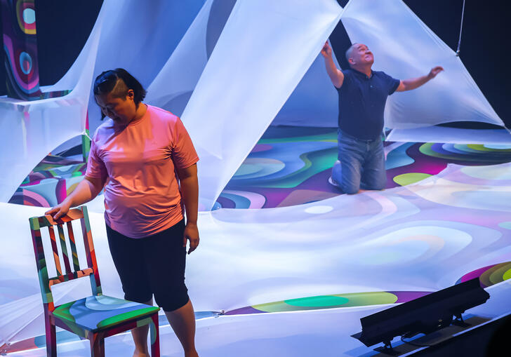 Two people perform on stage among large pieces of draping white fabric with bright lights projected onto it. 
