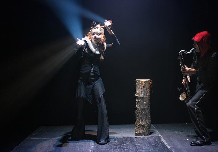 A person in a mask dances by a tree trunk while another person plays the saxophone. 