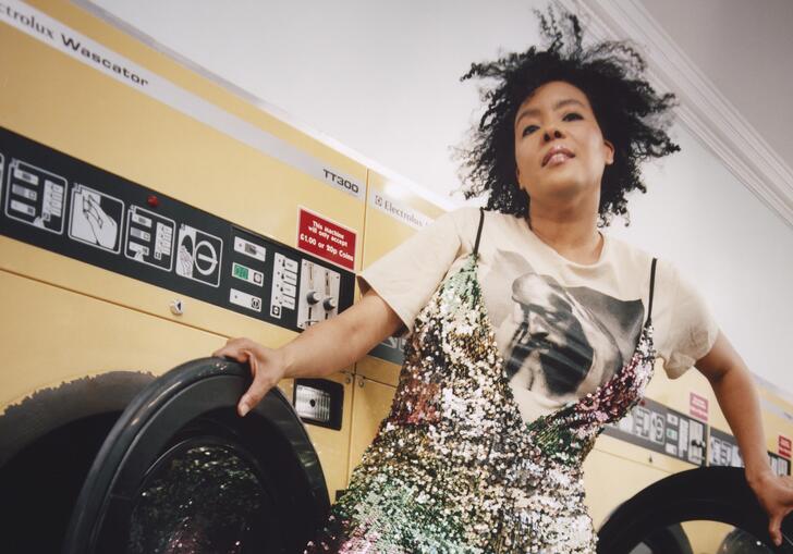 Elaine Mitchener standing in front of washing machines in a laundrette, holding the open washing machine doors.