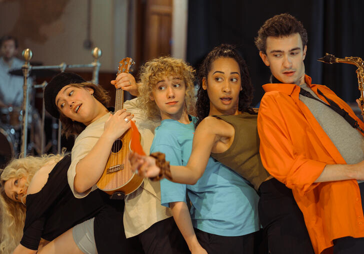 Five performers lean on each other in a line. Two play instruments.