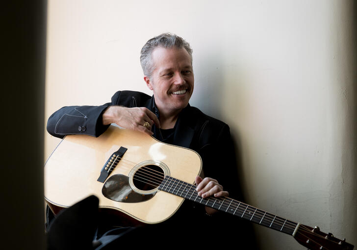 Jason Isbell smiling, sitting with guitar on lap
