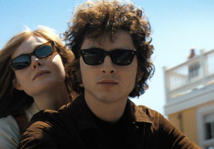Two people wearing sunglasses ride through an American town with a blue sky overhead.